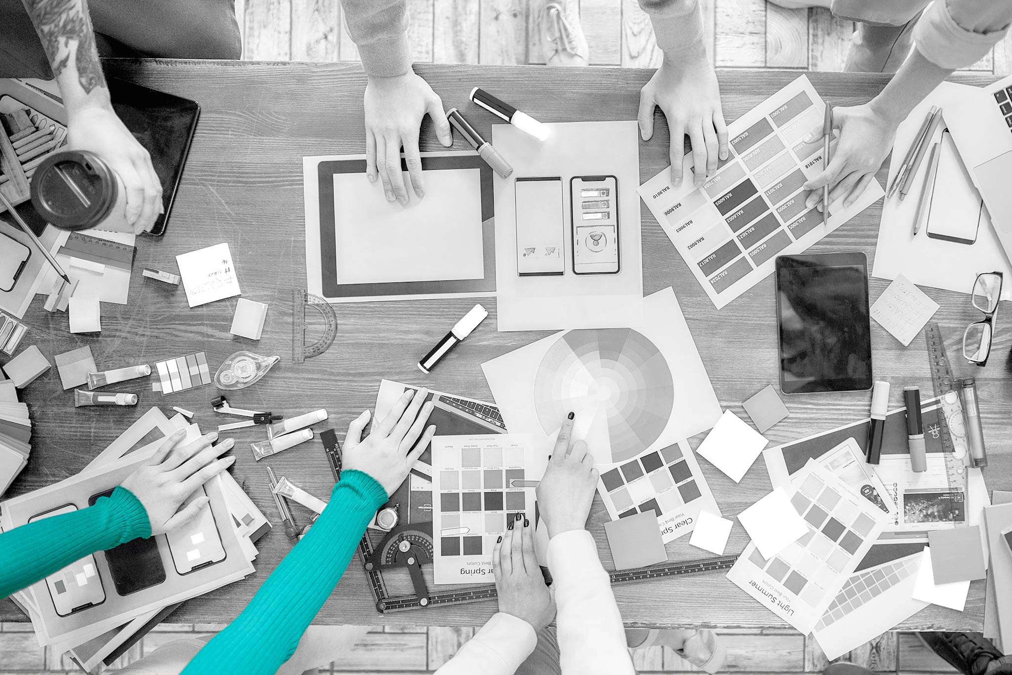 Black and white photo of a group of hands outstretched over a table with various design documents and color swatches, with one person's sweater being teal